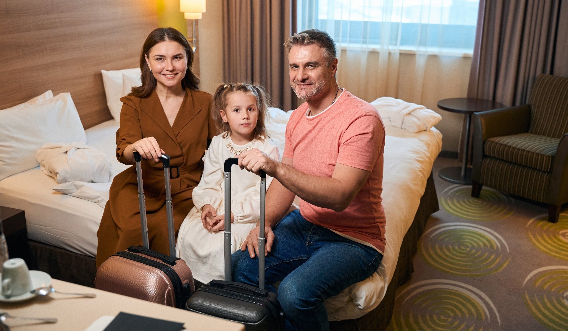 Family with suitcases sitting on bed and looking at camera in hotel room