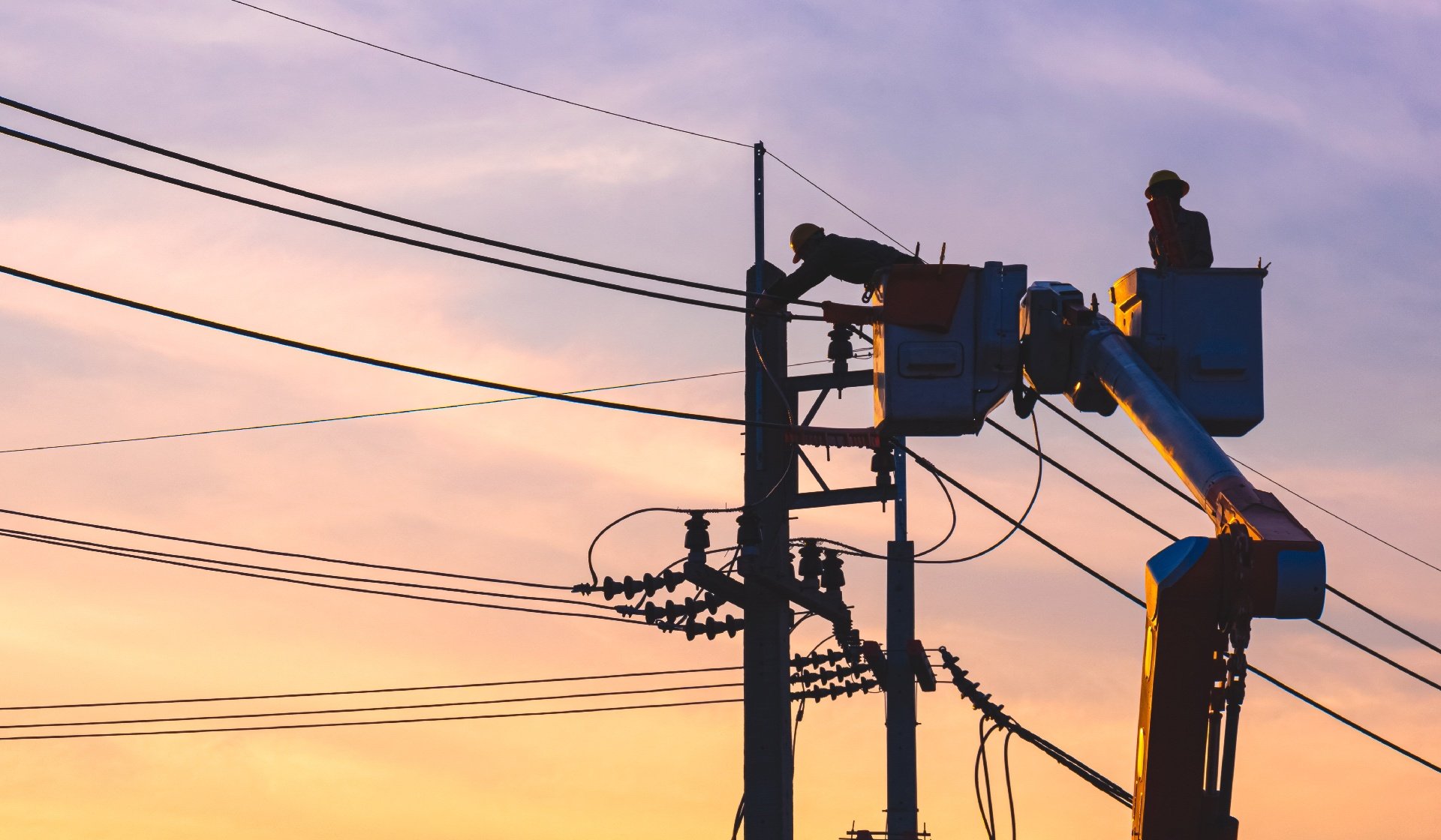 Silhouette 2 electricians in bucket boom truck repairing electrical system on power pole at sunset