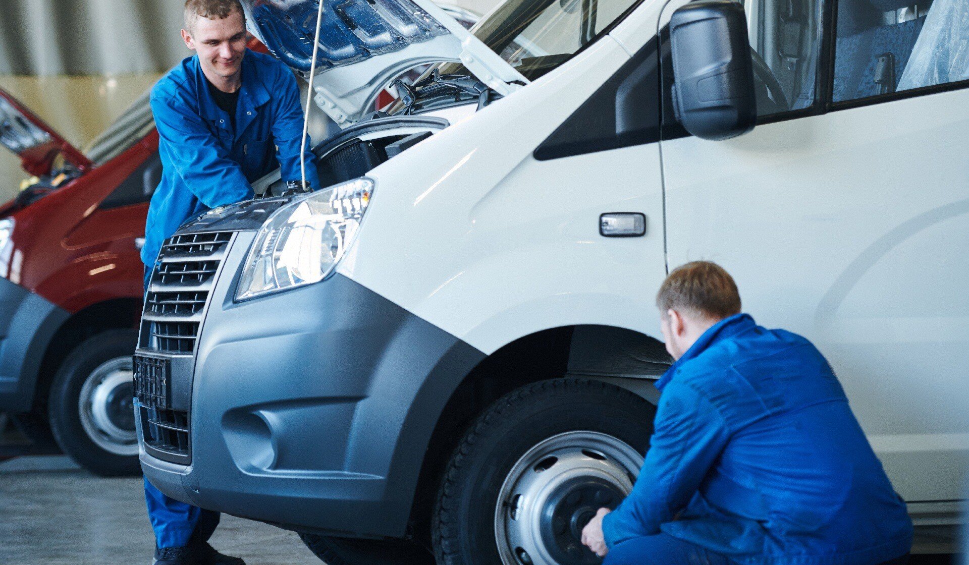 young mechanics in workwear repairing engine of car while his colleague checking its wheels in workshop